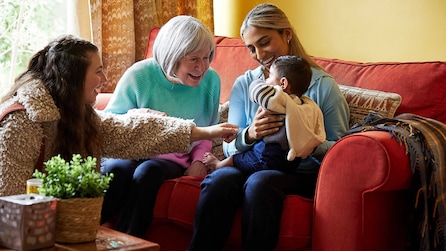 Maya holding a baby and sitting on a red couch next to Jackie and Ava 
