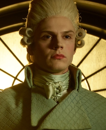 Evan Peters headshot wearing white baroque wig with silk white neck scarf and white coat in front of illuminated window