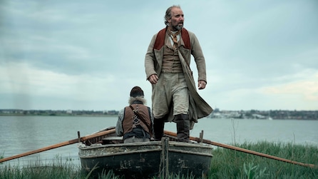 Two men in a boat placed on the grass with water in the background