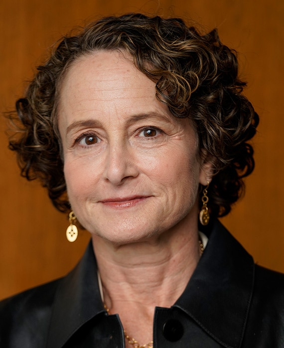 Headshot of Nina Jacobson wearing a black button up and gold jewelry, with a brown background behind her.