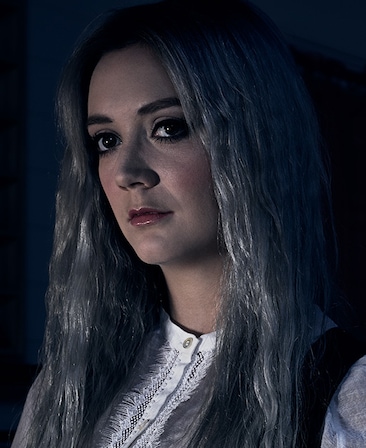 Billie Lourd headshot with long grey hair and smokey black liner in white lace button down in dark room with face illuminated