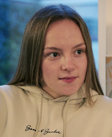Lili Jones wearing a tan pull over hoodie sitting in front of a window