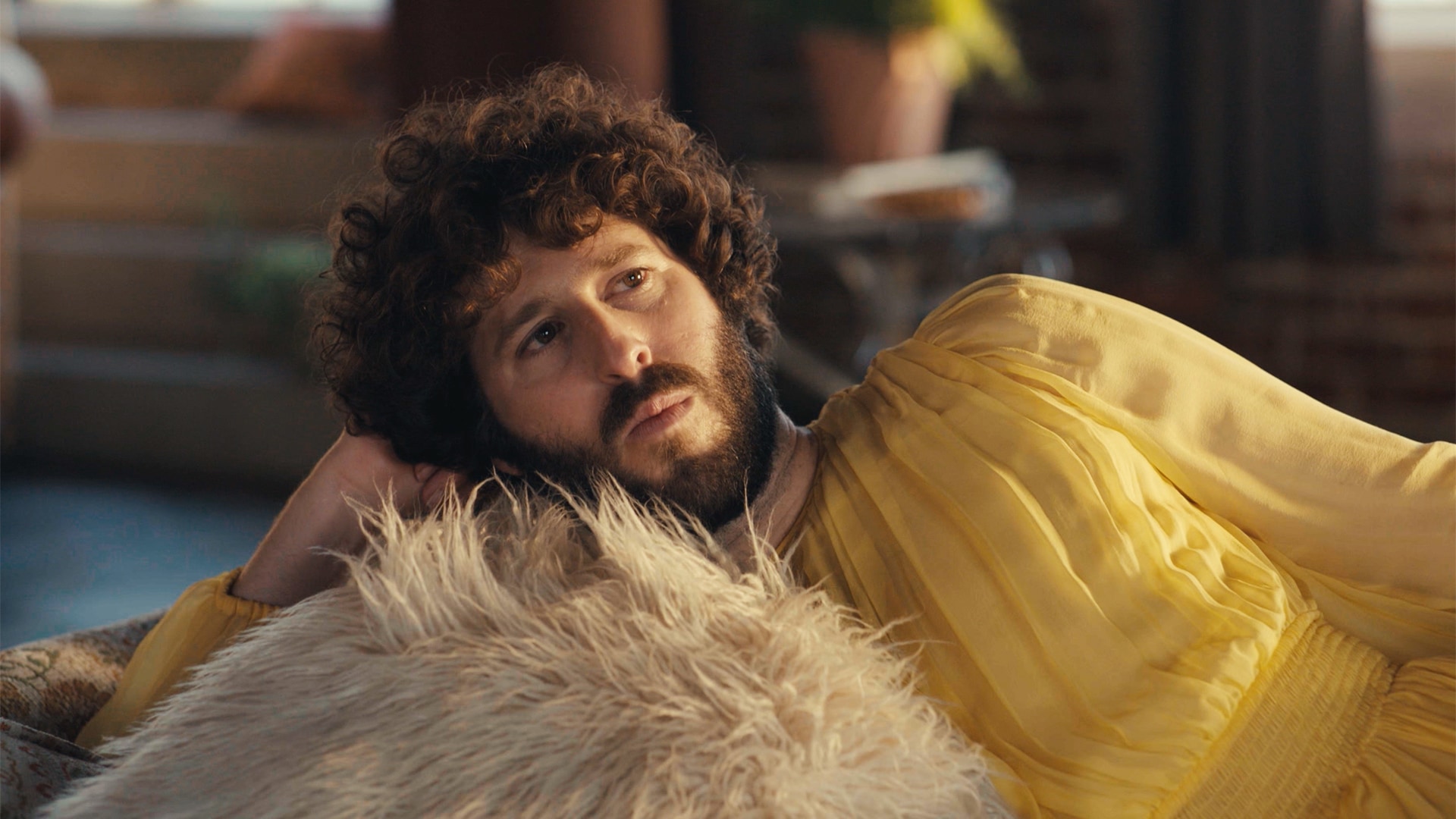 Dave Burd wearing a yellow dress and laying on a white fluffy pillow