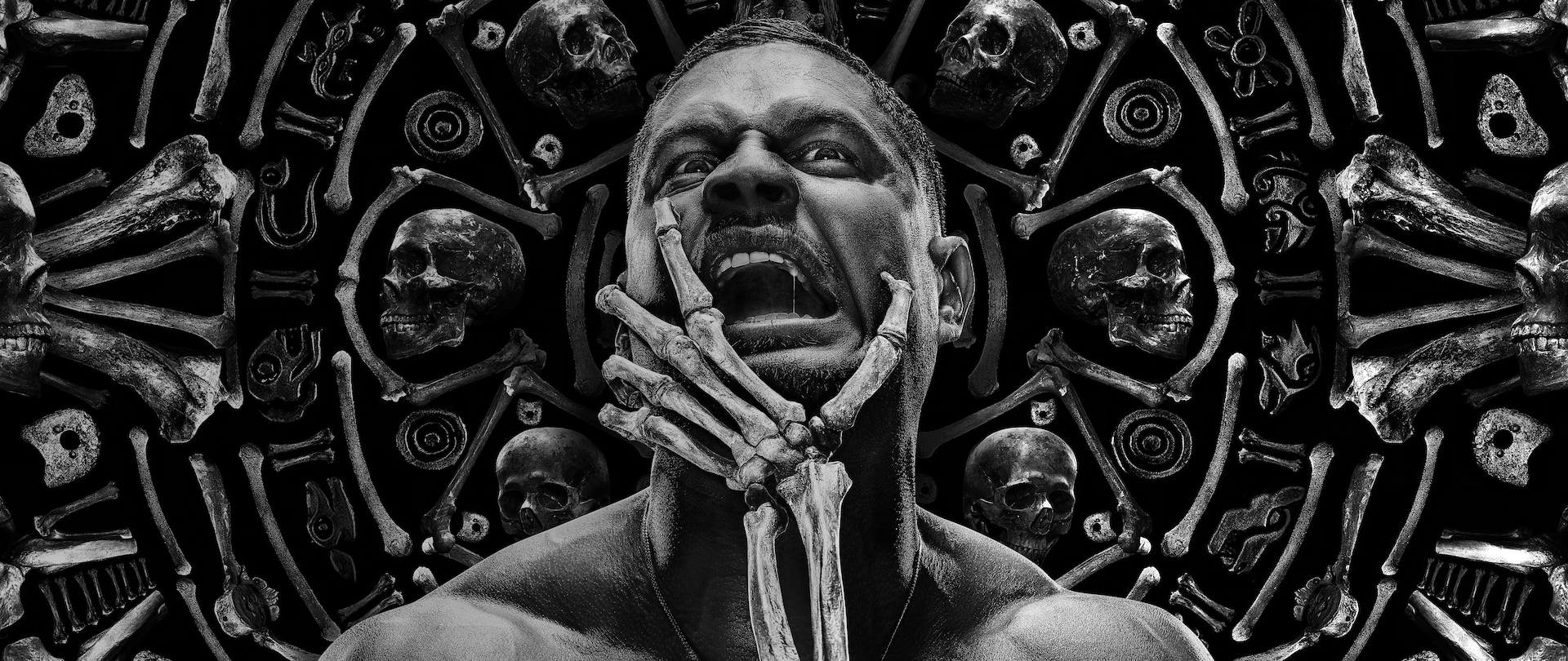 JD Pardo centered in front of bones motif in black and white with mouth open and a skeleton hand grabbing his chin