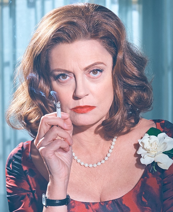 Susan Sarandon as Bette Davis smoking cigarette and wearing red and black scoop top and pinned white flower in Feud FX show