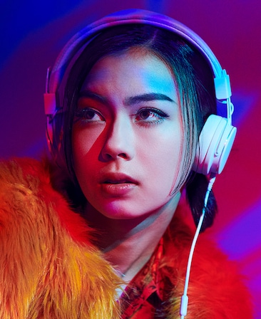 Lauren Tsai wearing white headphone in orange fur coat, looking up with neon purple and pink lighting over face and backgroun