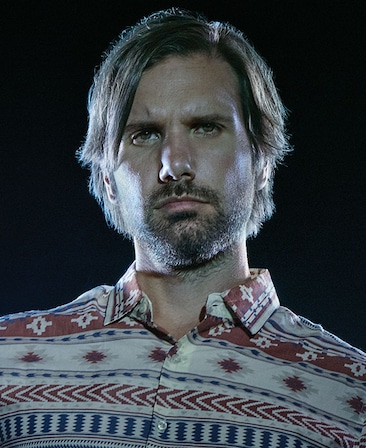 Jon LaJoie in red, white, and blue western style button down with plain black background and lights shining on face
