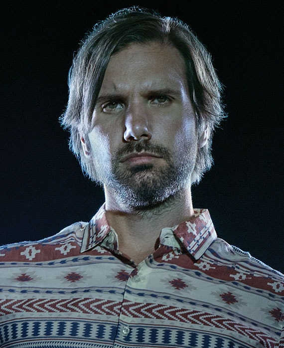 Jon LaJoie in red, white, and blue western style button down with plain black background and lights shining on face