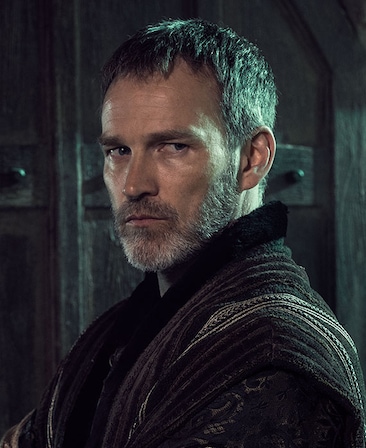 Stephen Moyer headshot in textured and gold detailed black jacket with ancient wood doors illuminated by moonlight behind him