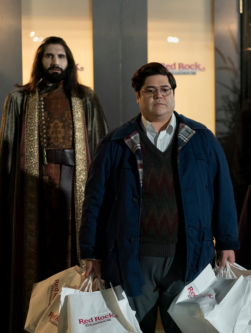 Guillermo holds bags while Nandor stands behind him. 
