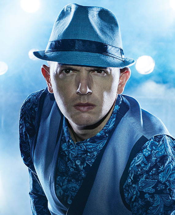 Paul Scheer headshot in blue western print top with satin blue vest, with blue fedora, with bright stadium lights behind him