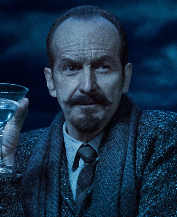 Headshot of Denis O'Hare with mustache in grey suit holding martini glass on cloudy night from FX's AHS Double Feature