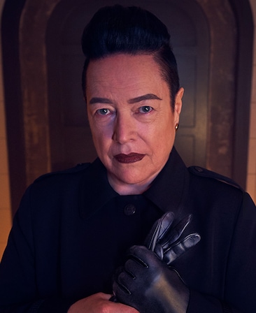 Kathy Bates headshot in black top holding black leather glove in front of wooden door from AHS Apocalypse