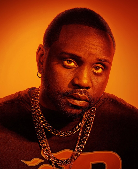 Headshot of Bryan Tyree Henry wearing thick silver chain necklaces with black shirt and heavy sunset lighting over face 