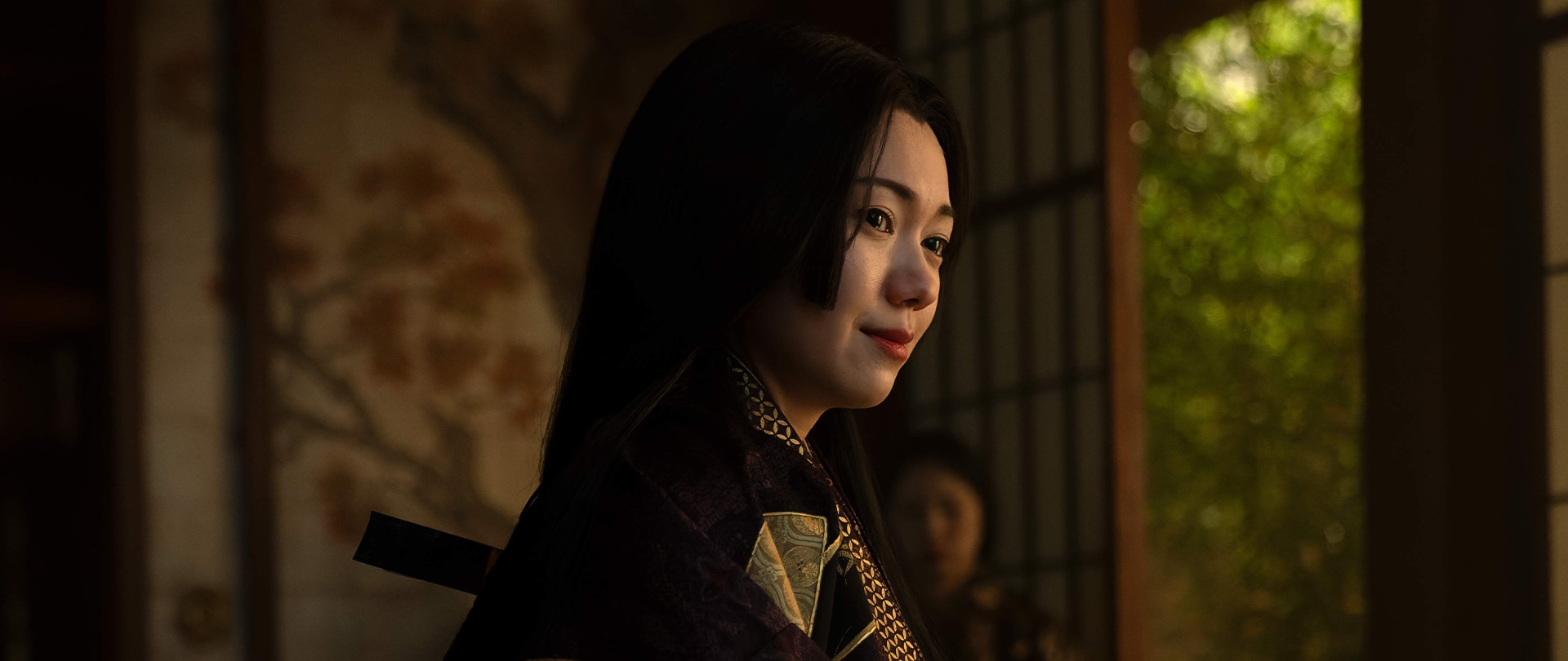 Ochiba sits in front of a low writing table dressed in a purple uchikake with gold fan patterns in FX’s Shogun