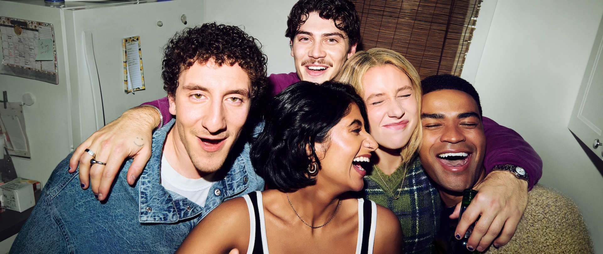 A group of five friends posing for a selfie while laughing in a small apartment kitchen.