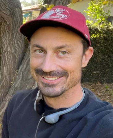 Headshot of executive producer Rob Rosell standing outside wearing a red baseball cap and gray headphone around neck 