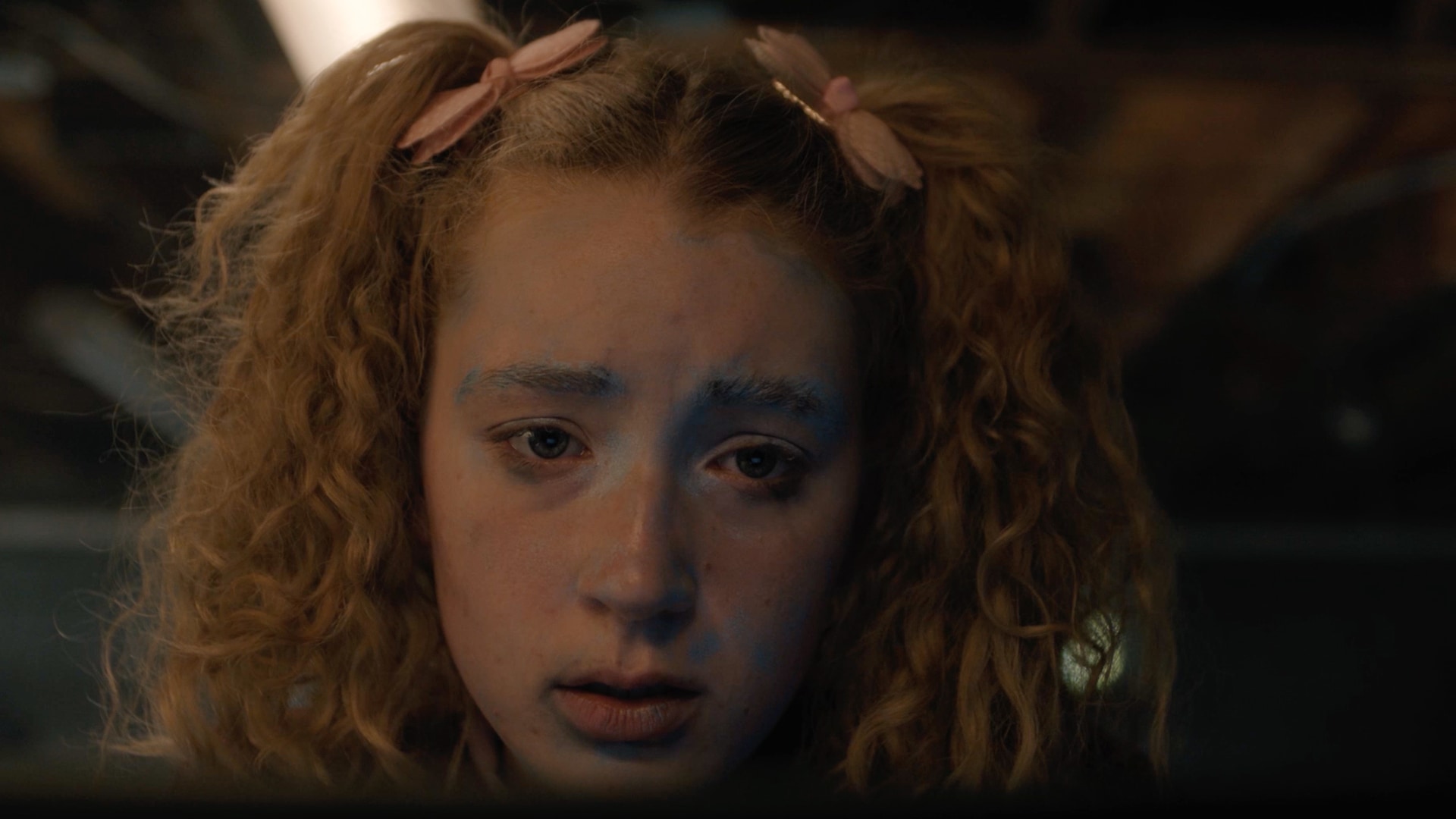 Girl with sparse blue face paint and blonde pigtails that have pink bows.