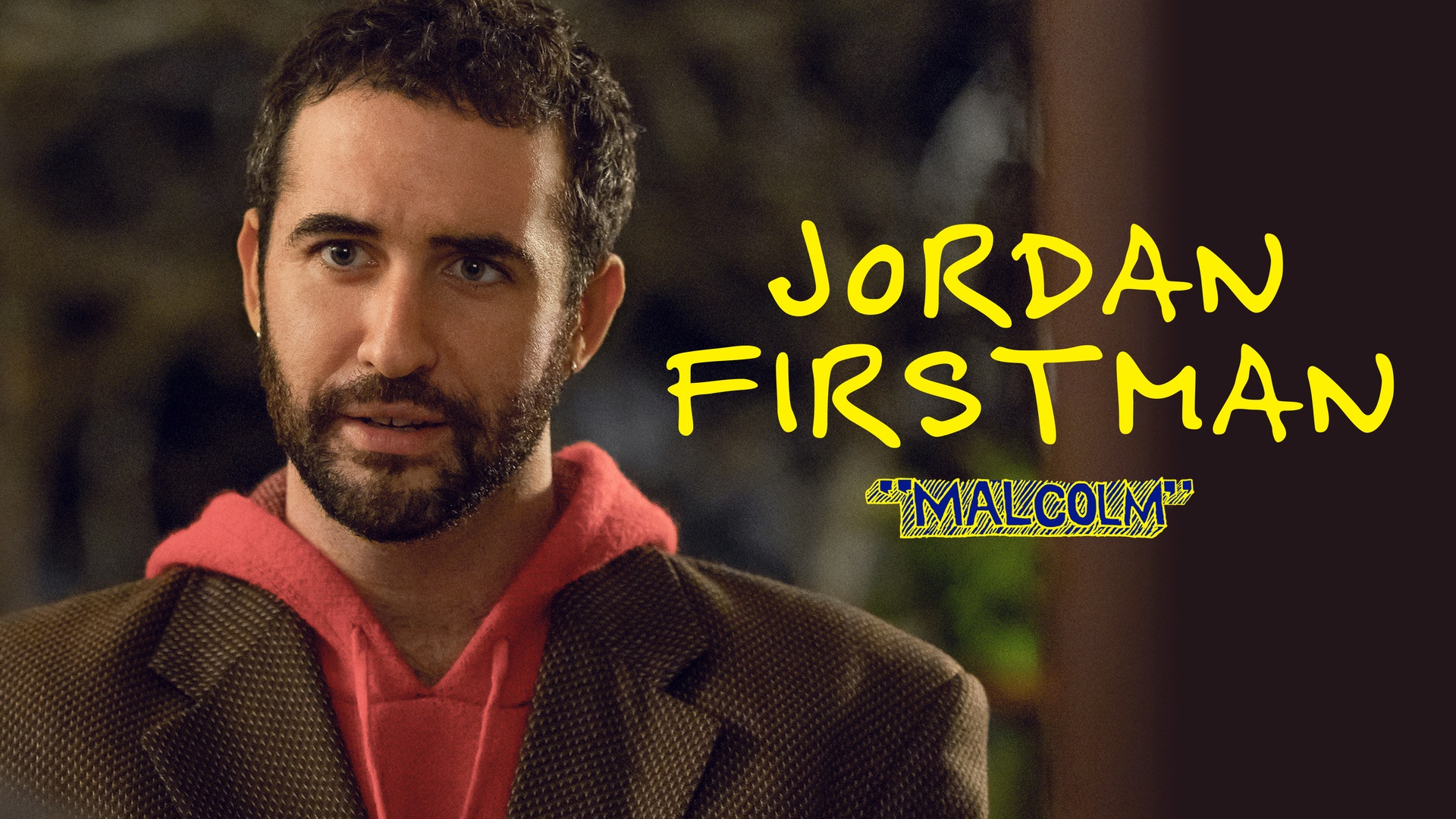 Jordan Firstman as Malcolm in FX's English Teacher, wearing a red hoodie over a brown pin stripped blazer..