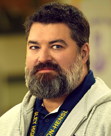 Sean Patton as Markie Hillridge for FX's English Teacher wearing a grey zip-up hoodie and blue collared shirt.