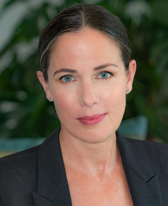 Headshot of Katherine Pope with slicked back hair, wearing a black suit, in front of greenery.