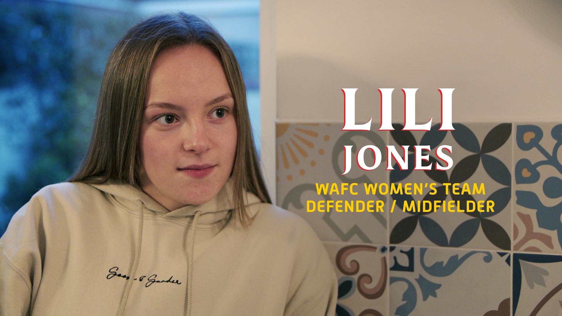Lili Jones wearing a tan pull over hoodie sitting in front of a window