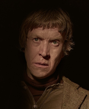 Sam Spruell headshot wearing a brown shirt and brown coat on top, standing in the dark
