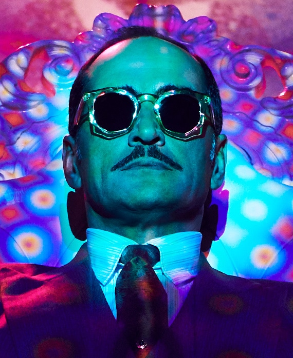Navid Negahban wearing angled sunglasses in suit with trippy neon purple lighting background with blue light shining on face