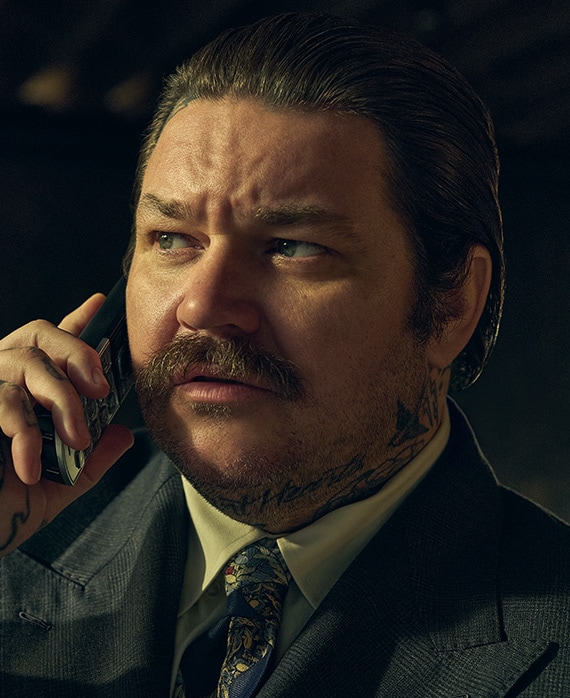 Matty Matheson headshot in FX's The Bear wearing a suit and tie, on the phone.