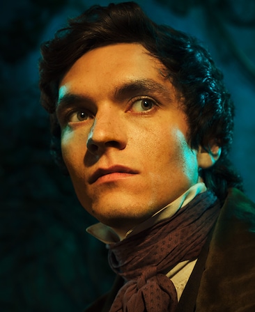 Fionn Whitehead headshot in black suede coat with black suit and brown neck tie scarf, with dark blue woods behind him