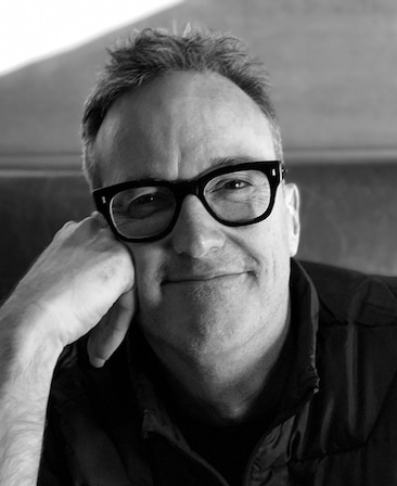 John Cameron black and white headshot in glasses andblack collared button down with right fist leaning on face