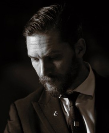 Tom Hardy black and white headshot in black suit and tie, looking down with soft light shining down on top part of face