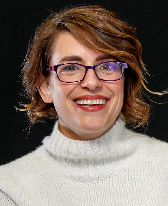 Anna Boden headshot wearing a white sweater and black glasses