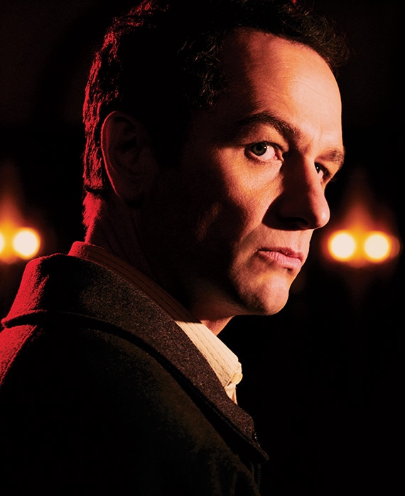 Matthew Rhys in black coat looking up over his right with half face illuminated at night with red lights in background