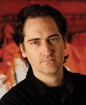 Daniel Zelman headshot in black jacket with red and white image background of stone sculptures