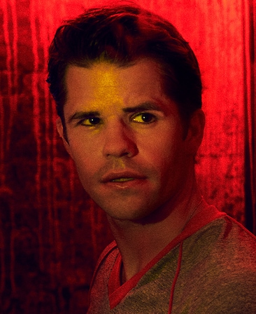 Headshot of Charlie Carver in grey shirt under red lighting