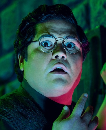 Headshot of Harvey Guillen as Guillermo wearing black glasses with pink and green lights shinning on his face.