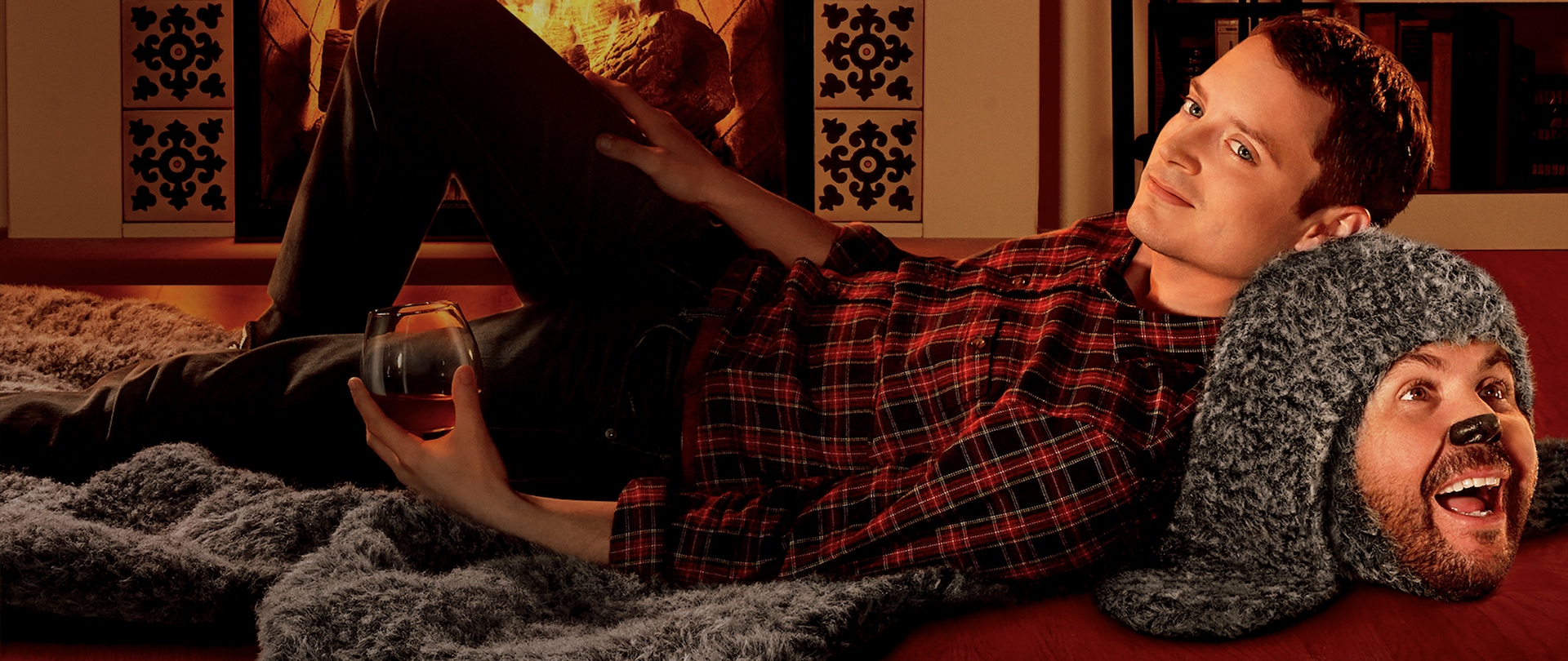 Elijah Wood lying in front of fireplace on top of grey "rug" of a man in a dog suit with his head popping up at the end front