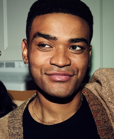 Headshot of Owen Thiele as Anton in FX's Adults wearing a black shirt and brown cardigan, wearing dainty gold jewelry.