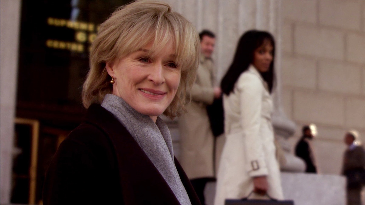 Patty Hewes wearing a black coat and gray scarf, standing outside a building