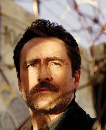 Demian Bichir on black jacket, standing in front metal barb wire gate, with sunlight and shadows on his face