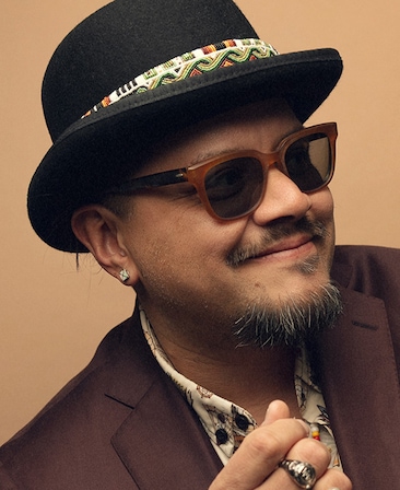 Headshot of executive producer Sterlin Harjo of FX's The Lowdown, wearing a brown suit, sunglasses and a black bowler hat.