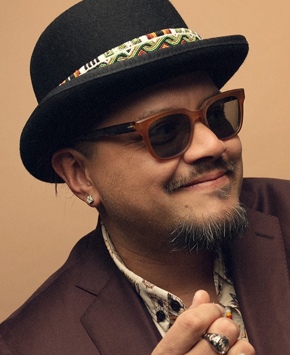 Headshot of executive producer Sterlin Harjo of FX's The Lowdown, wearing a brown suit, sunglasses and a black bowler hat.