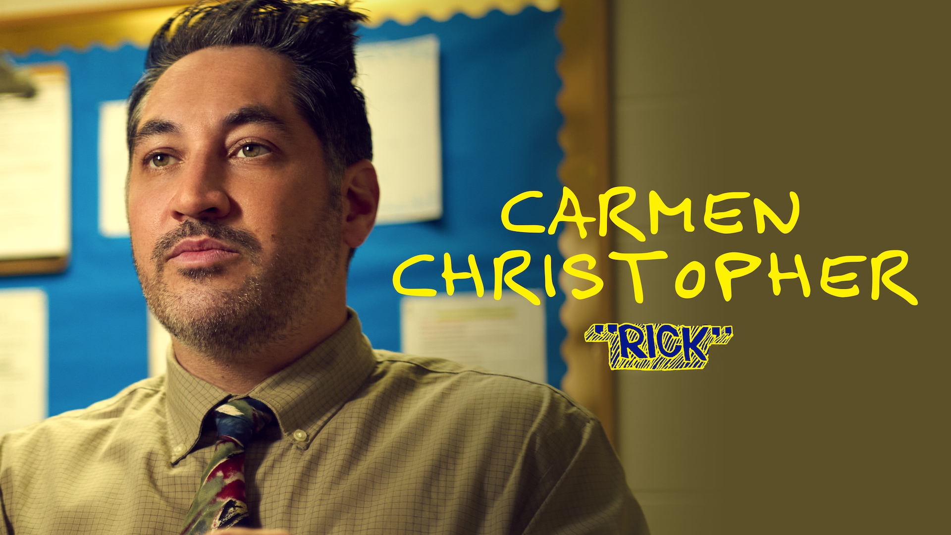 Carmen Christopher as Rick for FX's English Teacher wearing a green plaid button up and multi-colored tie facing to his side.