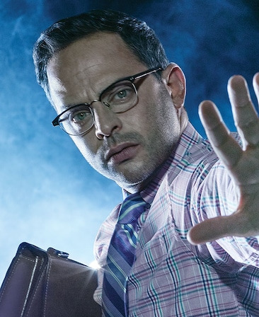 Nick Kroll in light purple plaid button down with striped purple tie, holding brown briefcase with left hand outstretched