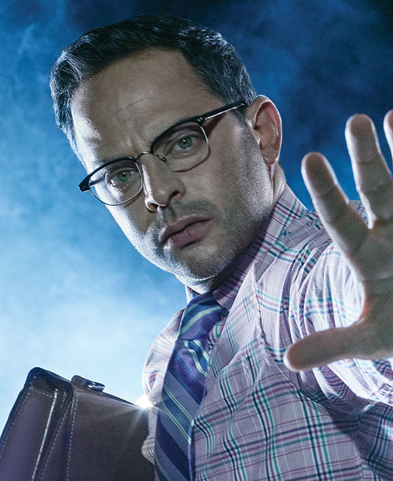 Nick Kroll in light purple plaid button down with striped purple tie, holding brown briefcase with left hand outstretched