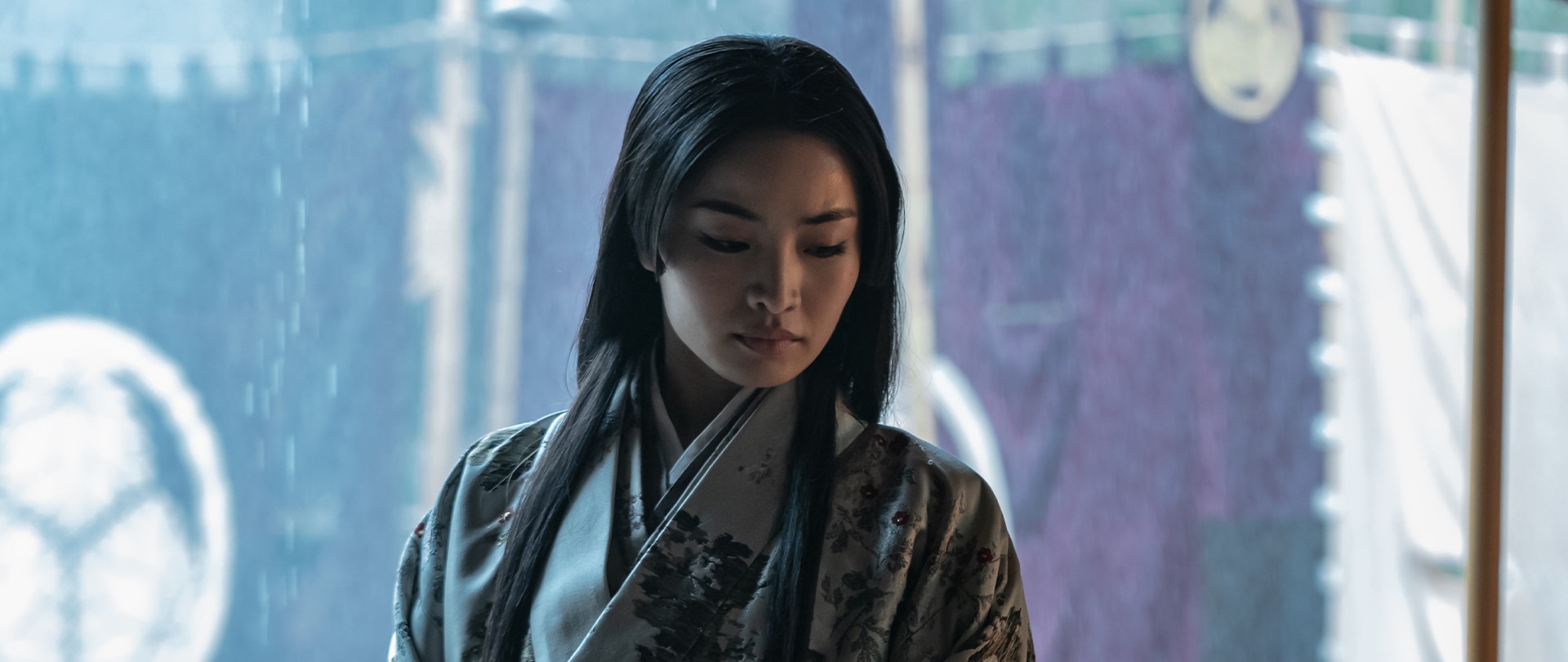Mariko gazes to the side wearing a light grey floral uchikake as rain pours outside Toranaga’s tent in FX’s Shogun