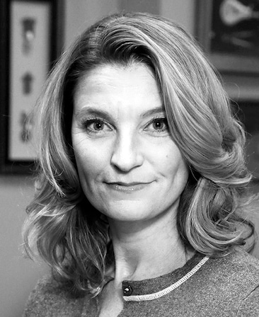 Black and white headshot of Meredith Stiehm with framed pictures and artwork behind her on wall