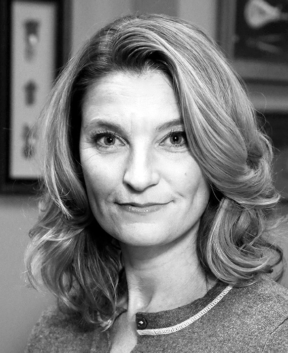 Black and white headshot of Meredith Stiehm with framed pictures and artwork behind her on wall