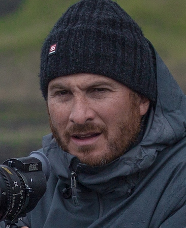 Darren Aronofsky headshot in dark grey parka and black beanie, holding specialized camera lens with green hill background